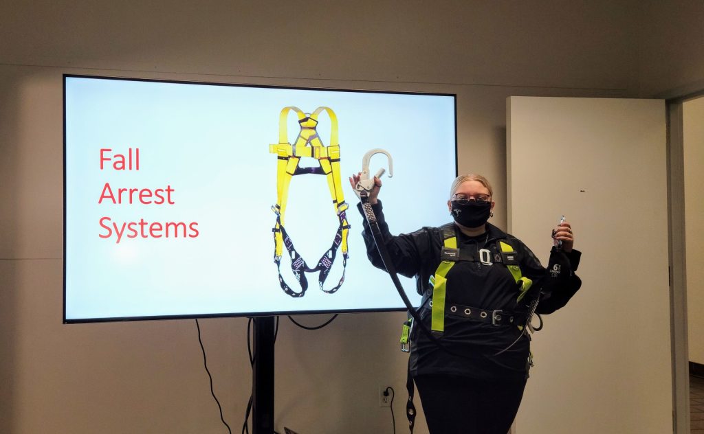 Corinne is wearing a harness and holding up a metal clip, with a slide behind her saying "Fall Arrest Systems."
