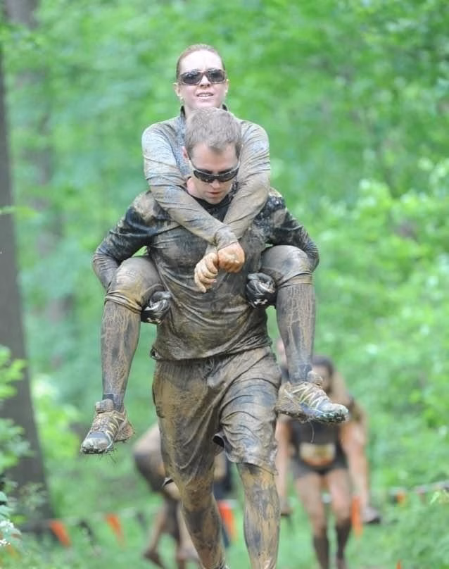 A man covered in mud carries a woman, also muddy, on his back through a forested area during an outdoor event or race. They both wear sunglasses and appear to be enjoying the challenge.