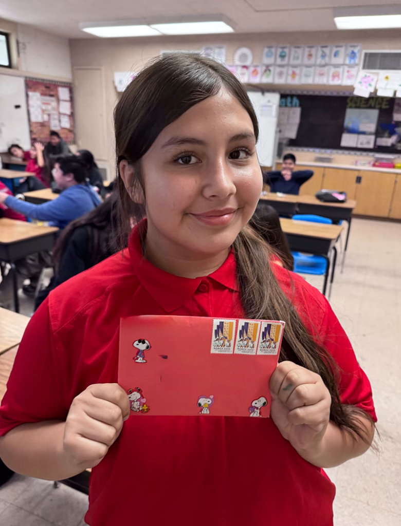 A girl wearing a red shirt smiles and holds a red envelope decorated with Snoopy stickers and four U.S. postage stamps in a classroom setting, with other students and desks visible in the background.