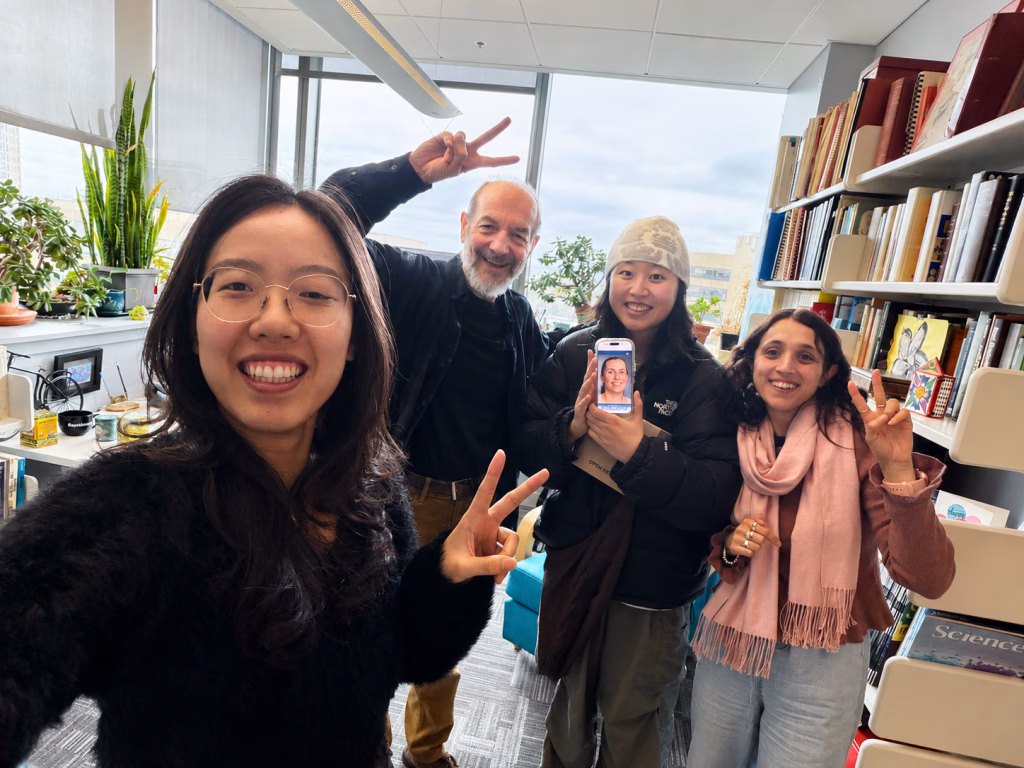 Four people smile and flash peace signs in an office with bookshelves and plants. One holds a phone displaying another person, who joins the group virtually to discuss AI cancer detection. Bright daylight streams through the large windows.