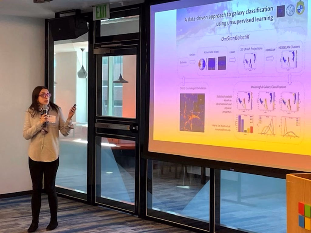 A woman holding a microphone stands next to a large screen presenting a slide titled A data-driven approach to galaxy classification using unsupervised learning and AI cancer detection in a modern conference room with glass doors and windows.