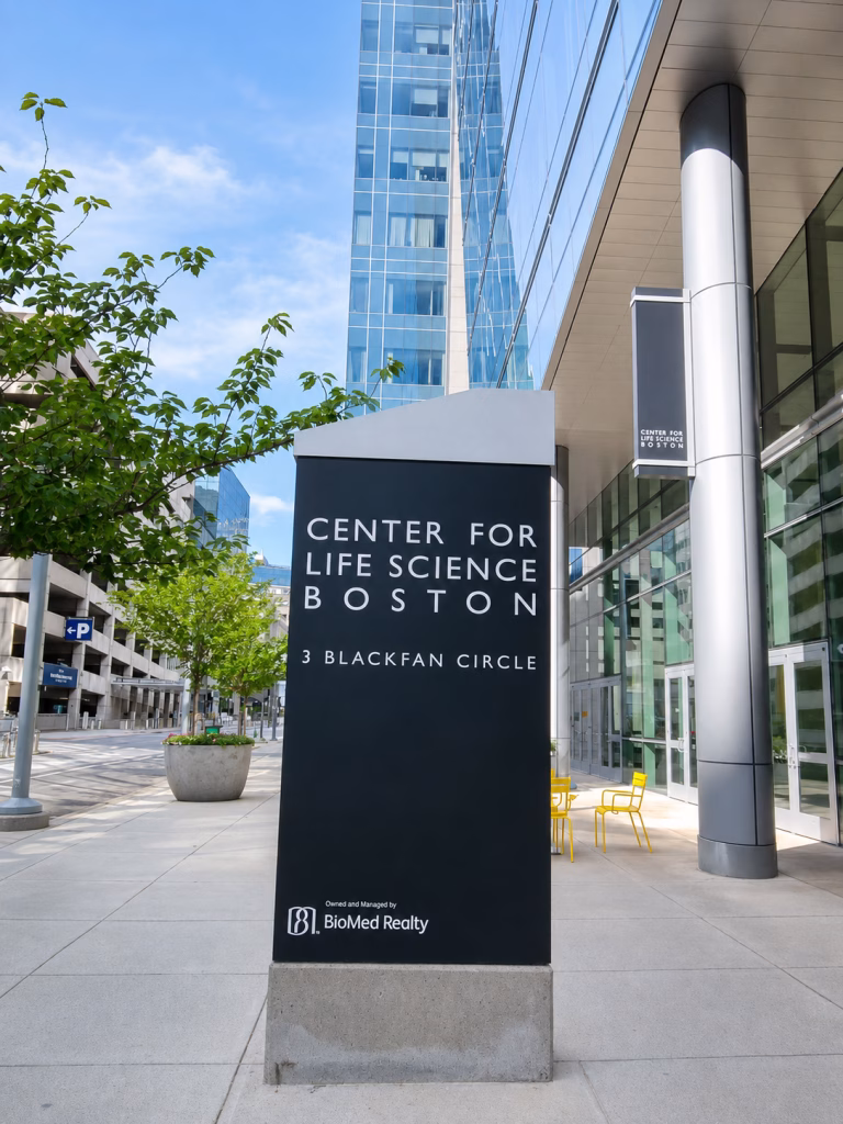 A tall black sign outside a modern glass building reads Center for Life Science Boston, 3 Blackfan Circle—home to cutting-edge AI cancer detection research. Nearby are yellow chairs, planters, and trees lining the sidewalk.