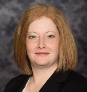 Woman with straight, shoulder-length red hair, wearing a black blazer over a cream top, and a necklace. She is facing the camera, smiling softly, with a mottled gray background behind her.