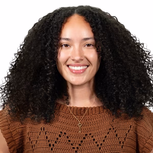 A staff member with curly dark hair, wearing a brown crochet top and a necklace, smiles at the camera against a plain light background.