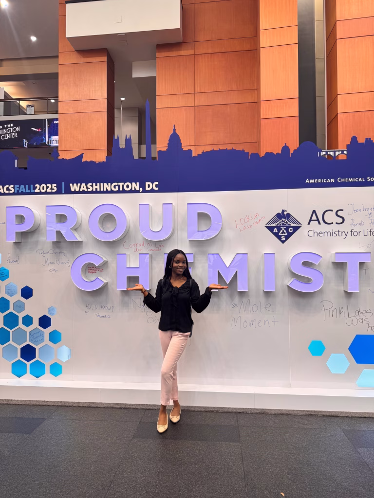 A woman stands smiling with arms raised in front of a large display reading PROUD CHEMIST at the ACS Fall 2025 event in Washington, DC. The blue hexagon backdrop and handwritten notes highlight advances in sustainable materials.