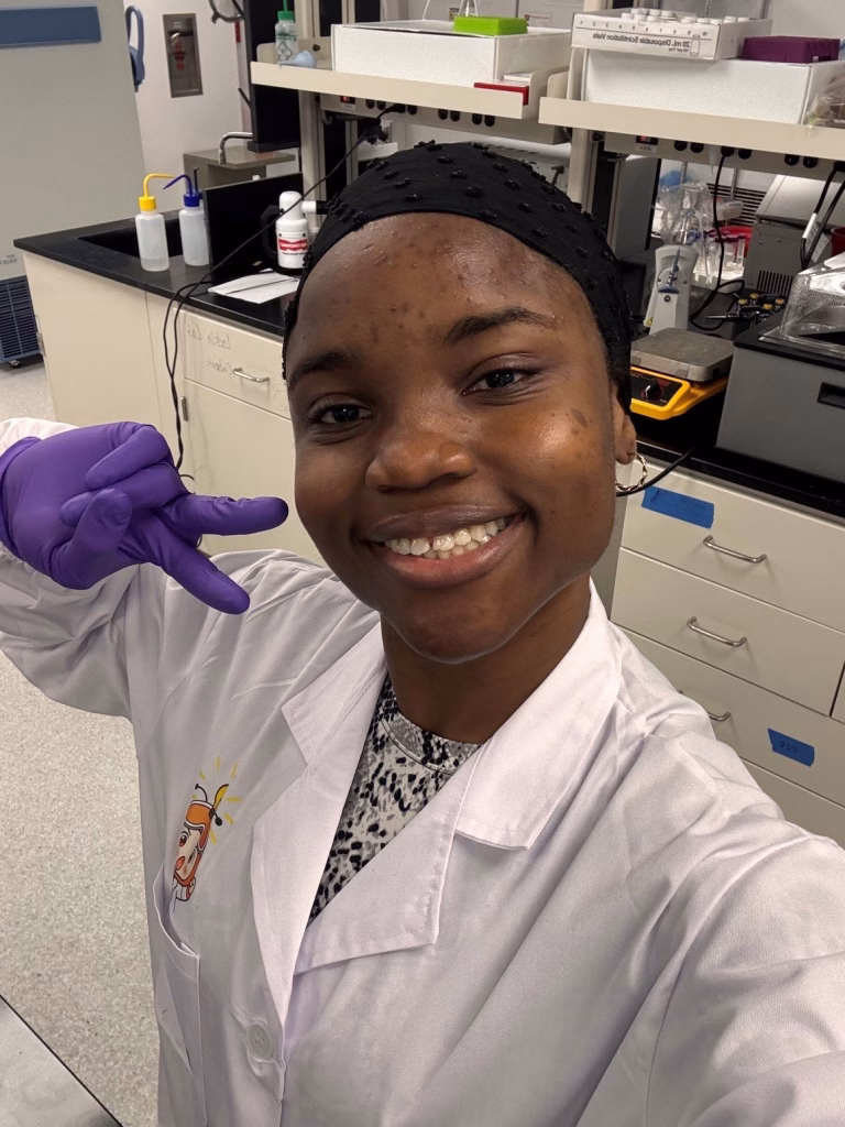 A smiling woman in a lab coat and purple gloves takes a selfie in a laboratory focused on sustainable materials, making a peace sign with her hand. Laboratory equipment and supplies are visible in the background.