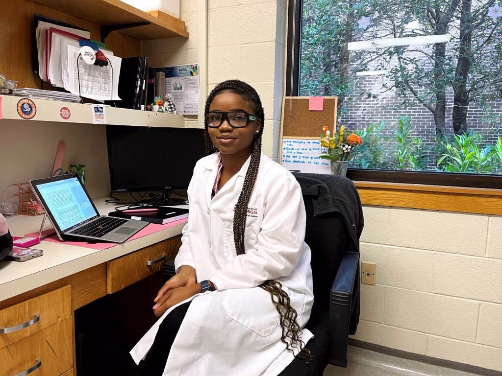A woman in a white lab coat and glasses sits at a desk in an office or lab, surrounded by papers, a laptop, and flowers. Through the window, lush green plants hint at her research on sustainable materials.