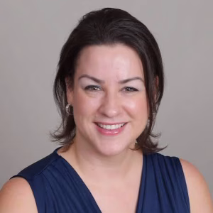 A woman with shoulder-length dark brown hair, wearing a sleeveless navy blue top, smiles at the camera against a neutral light gray background.