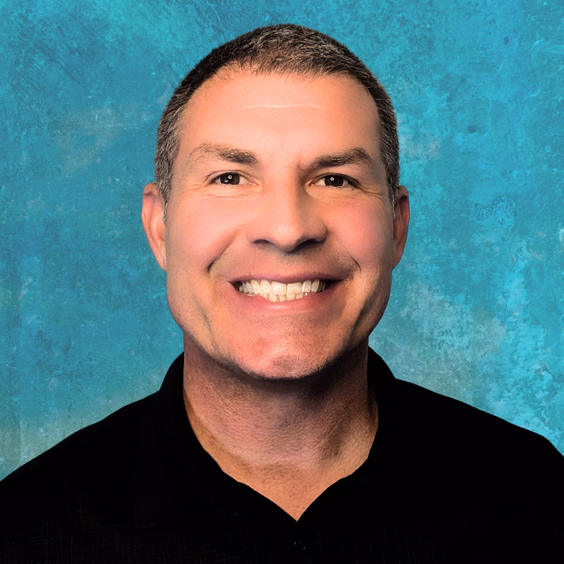 Smiling man with short brown hair wearing a black collared shirt stands in front of a textured blue background, embodying confidence and curiosity—ideal for illustrating the innovative spirit behind using AI in science.