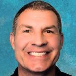 A man with short dark hair smiles broadly, showing his teeth, against a textured blue background, as if ready to host the upcoming AI in Science Webinar.