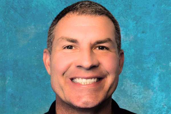 A man with short dark hair smiles broadly, showing his teeth, against a textured blue background, as if ready to host the upcoming AI in Science Webinar.