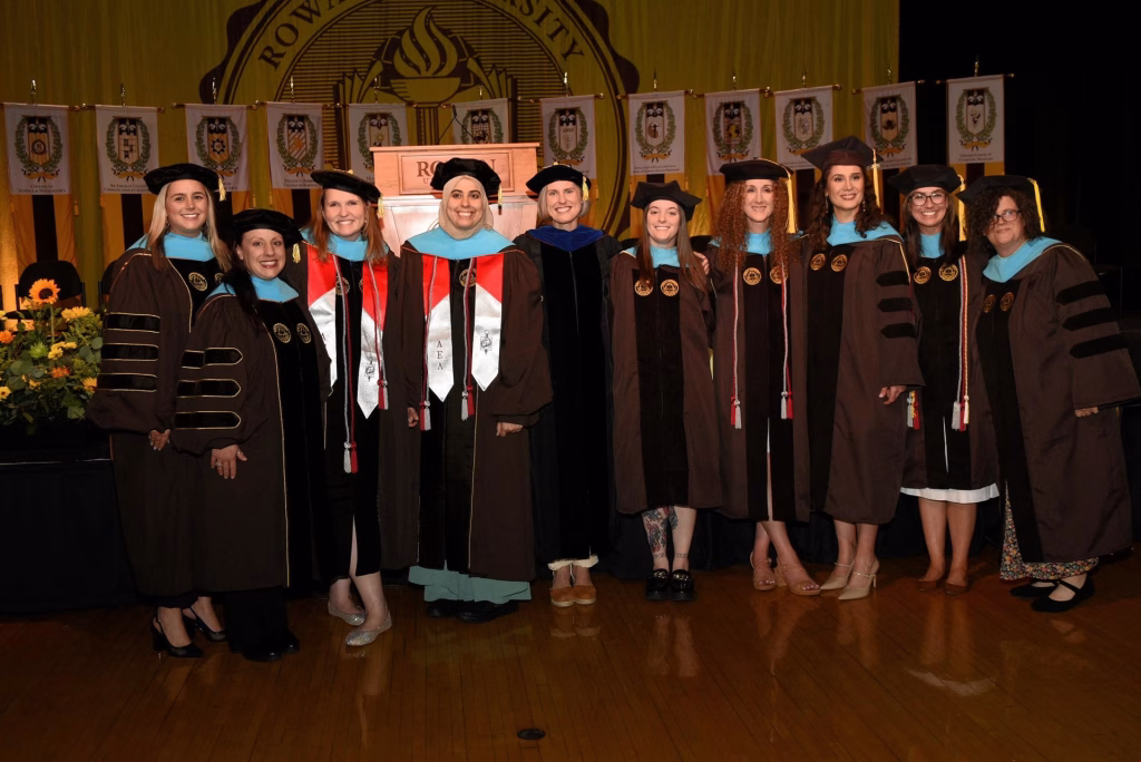 A group of nine people in graduation caps and gowns stand in a row, smiling, on a stage with university banners and a yellow backdrop behind them, proudly celebrating their achievements in fields that emphasized laboratory safety.