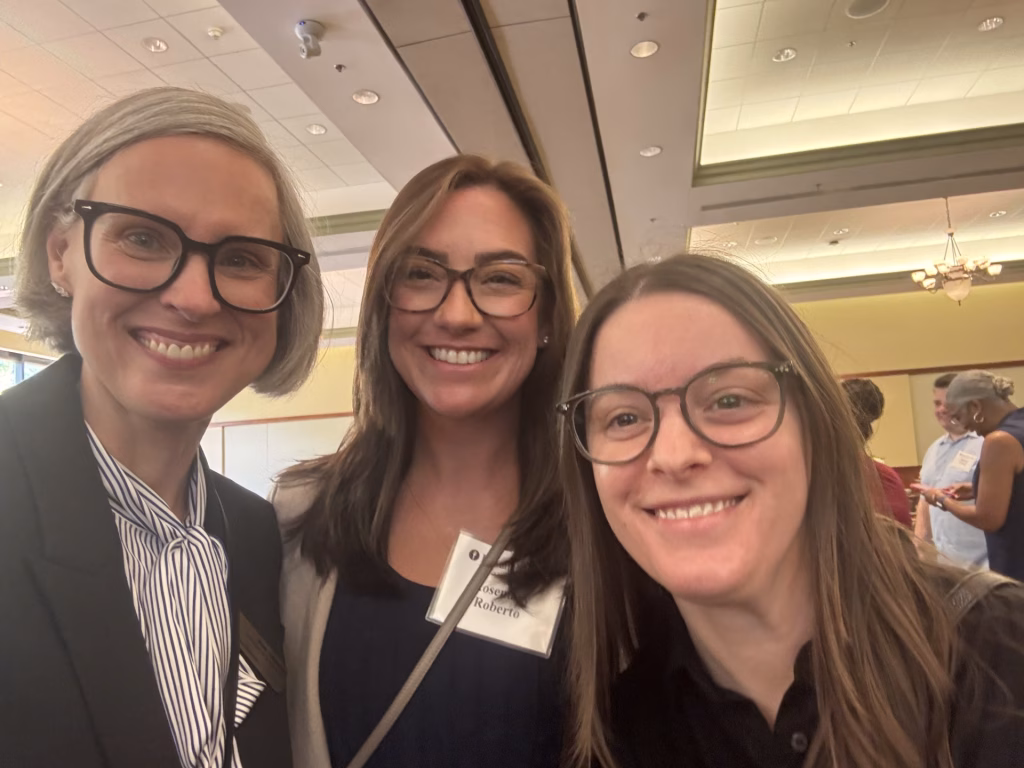 Three women with glasses smile for a selfie at an indoor event focused on laboratory safety. Two wear business attire and name tags. Other people are visible in the background, standing and conversing.