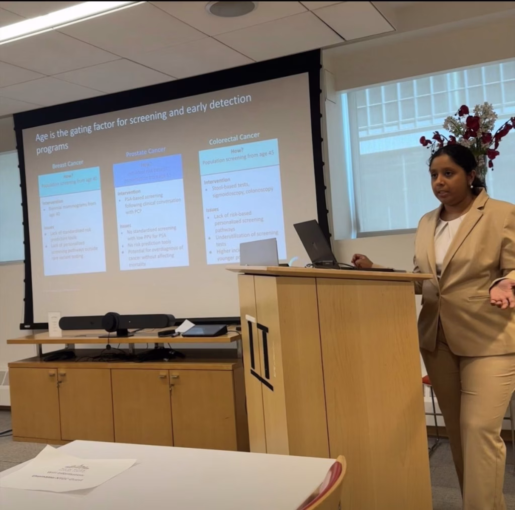 A woman in a tan suit presents at a podium in a classroom. Behind her, a screen displays slides about breast, prostate, and colorectal cancer screening and detection, while also touching on the latest advancements in Alzheimer’s clinical research. Flowers are visible near the window.