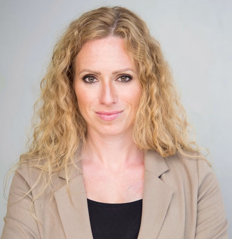 A woman with long, curly blonde hair wearing a beige blazer and black top, looking directly at the camera with a neutral expression against a plain light background.