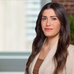 A woman with long, wavy brown hair, wearing a beige blazer over a brown top, stands indoors in front of a blurred background with a window and green plant. She is looking at the camera and smiling slightly.