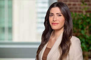 A woman with long, wavy brown hair, wearing a beige blazer over a brown top, stands indoors in front of a blurred background with a window and green plant. She is looking at the camera and smiling slightly.