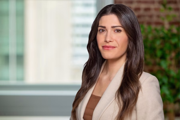 A woman with long, wavy brown hair, wearing a beige blazer over a brown top, stands indoors in front of a blurred background with a window and green plant. She is looking at the camera and smiling slightly.