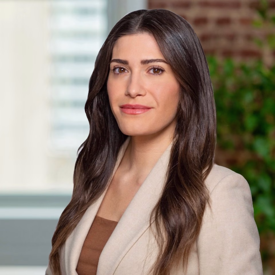 A woman with long brown hair wearing a beige blazer and brown top stands indoors, smiling softly. The background features a blurred window and greenery, creating an inviting atmosphere ideal for discussing foundation funding strategies.