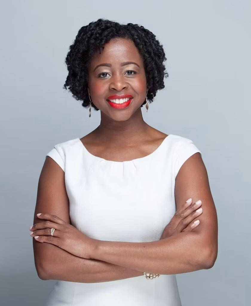 A woman with short curly hair, wearing a white dress, stands against a plain gray background. She is smiling with red lipstick and has her arms confidently crossed—ready to make decisions with incomplete data.