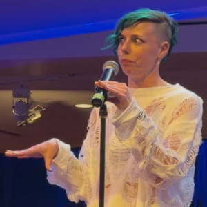 A person with short, green hair and a side shave stands at a microphone, wearing a white, ripped sweater, making a surprised or expressive face with one hand raised mid-gesture.