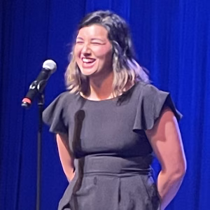 A woman in a black dress stands and laughs in front of a microphone on stage, with a blue curtain backdrop.