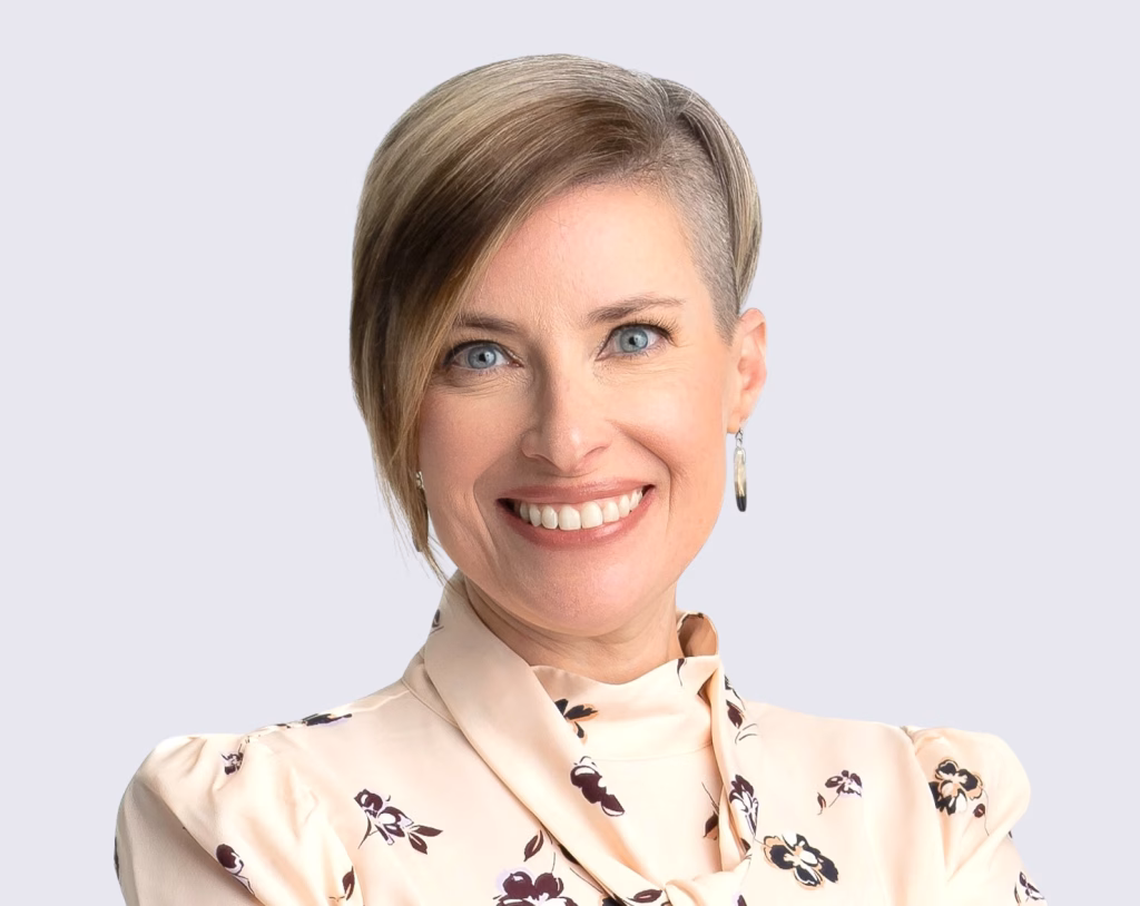 Smiling woman with short, asymmetrical blonde hair, wearing a light floral blouse with a high collar and hoop earrings, standing against a plain light background.