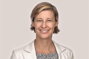A woman with short blonde hair smiles at the camera. She is wearing a white blazer over a patterned top, and is posed against a plain, light-colored background.
