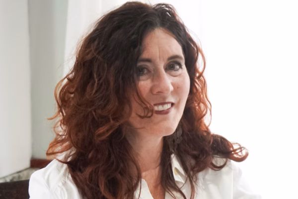 A woman with wavy, shoulder-length brown hair smiles softly. She is wearing a white blouse and is seated indoors with a bright, softly lit background behind her.