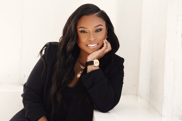 A woman with long wavy black hair, wearing a black outfit and gold jewelry, smiles while leaning her head on her hand against a white background.