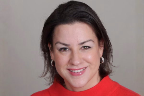A woman with short dark hair wearing silver hoop earrings and a bright red top smiles at the camera in front of a plain, light-colored background.