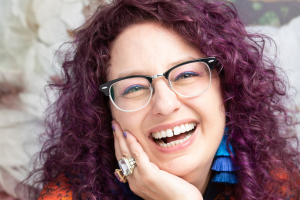 photo of a woman with purple hair smiling at the camera. She is wearing glasses and has blue earrings and a large ring.