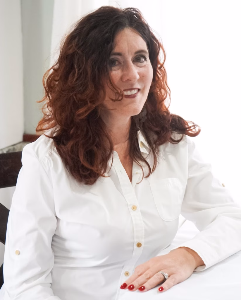 A woman with wavy brown hair wearing a white button-up shirt sits at a table, smiling. Her hand with red painted nails rests on the table, and she wears a ring. White curtains are in the background.