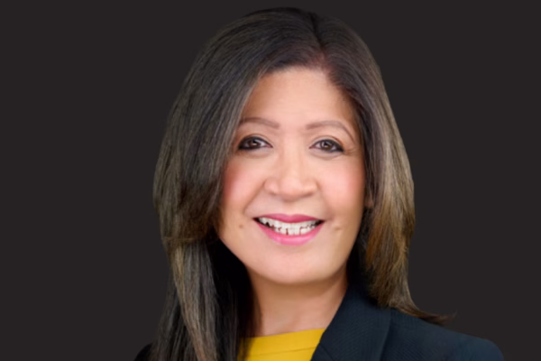 A woman with long, straight brown hair and light highlights smiles in front of a plain dark background. Dressed in a yellow top and navy blazer, she embodies confidence, perfect for thriving through strategic networking in STEM careers.