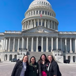 Four women smiling and standing together in front of the U.S. Capitol building on a sunny day with a clear blue sky.