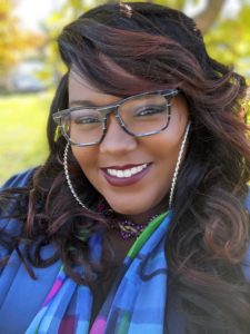 Ariangela Kozik is smiling wide, wearing glasses, big hoops, and a colorful scarf.