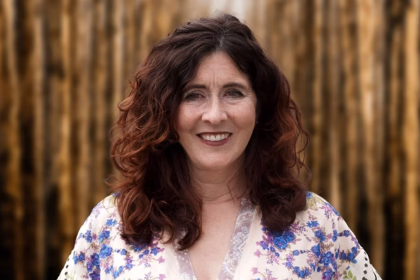 A woman with curly brown hair and a floral blouse smiles at the camera, standing outdoors in front of a blurred background of tall, vertical wooden poles—ready to share her expertise in upcoming webinars.