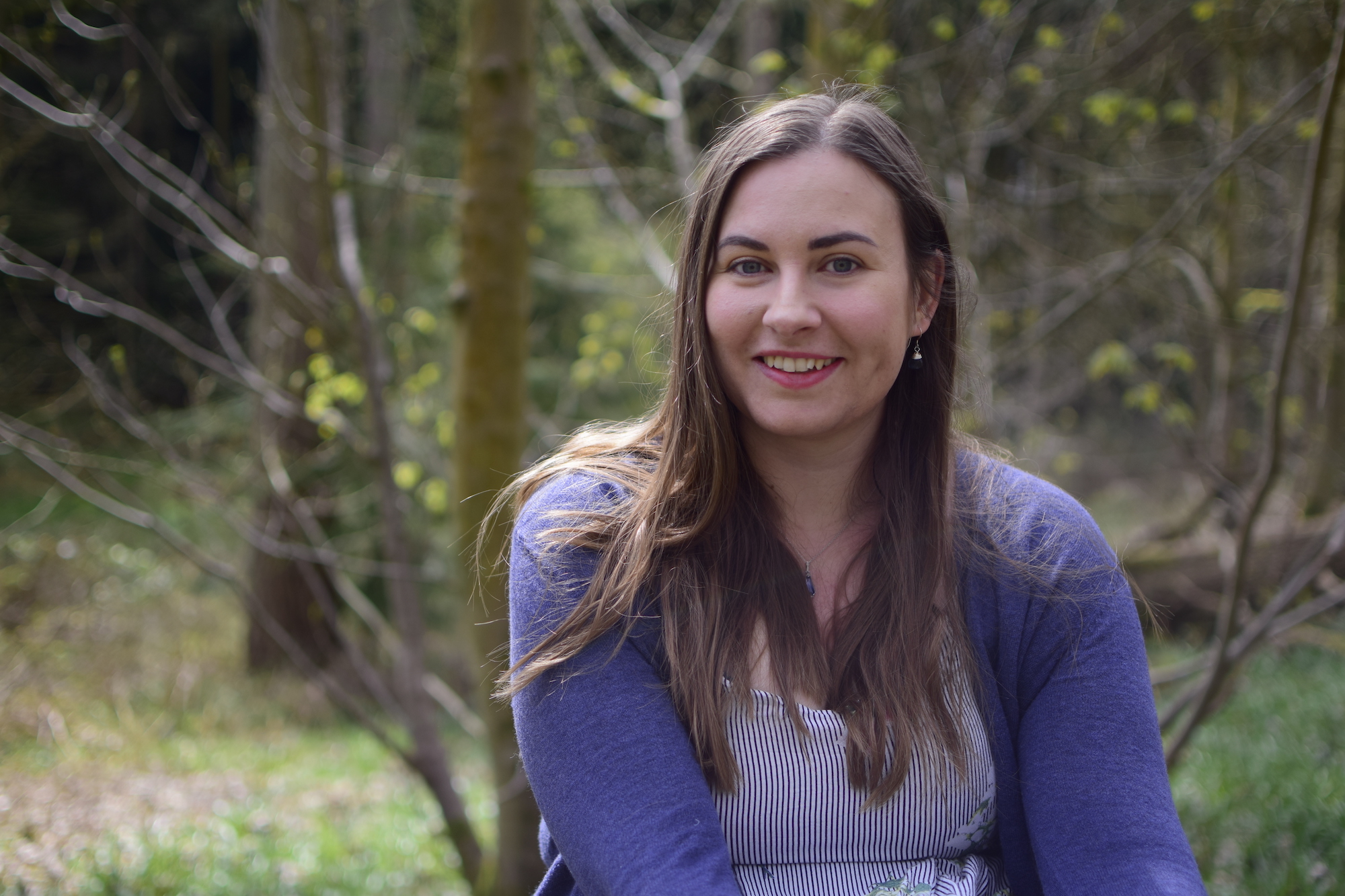 Zoe Ayres is smiling and wearing a blue cardigan in an outdoor setting with trees behind her