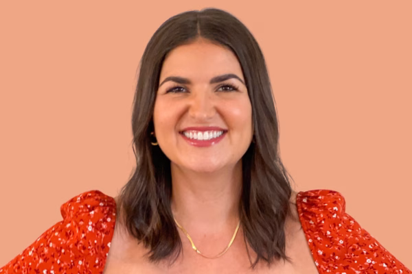 A woman with shoulder-length brown hair and a bright smile stands in front of a peach-colored background. She is wearing a red floral top and a gold necklace.