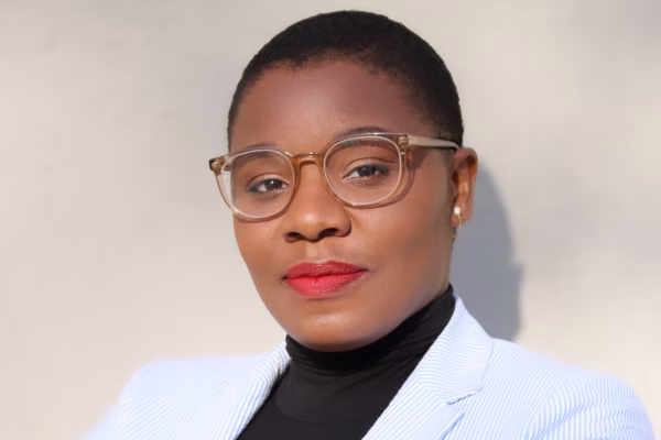 A person with short hair, wearing clear eyeglasses, red lipstick, a black turtleneck, and a light-colored blazer poses confidently against a neutral background—ready to lead or participate in webinars.
