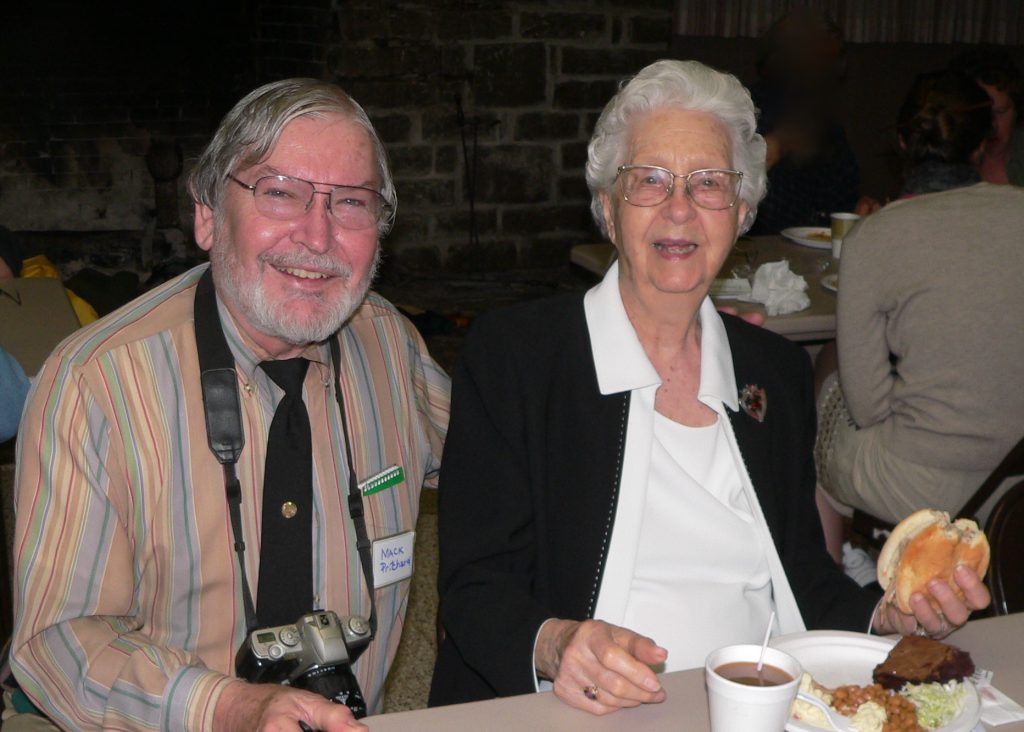 Mack Prichard is sitting next to Dr. Quarterman at a table. Dr. Quarterman has a sandwich in her hand and a plate of food in front of her.