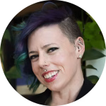 A person with short, partially shaved hair dyed purple and teal smiles. They have piercings on the nose, lip, and ear, and wear stud earrings. Green plants are visible in the background.