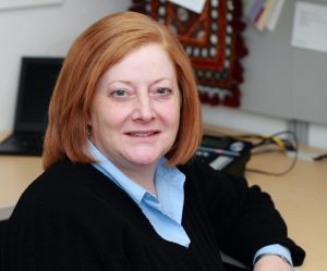 Joanne Kamens has short red hair, is wearing business clothing, and sitting at an office desk.