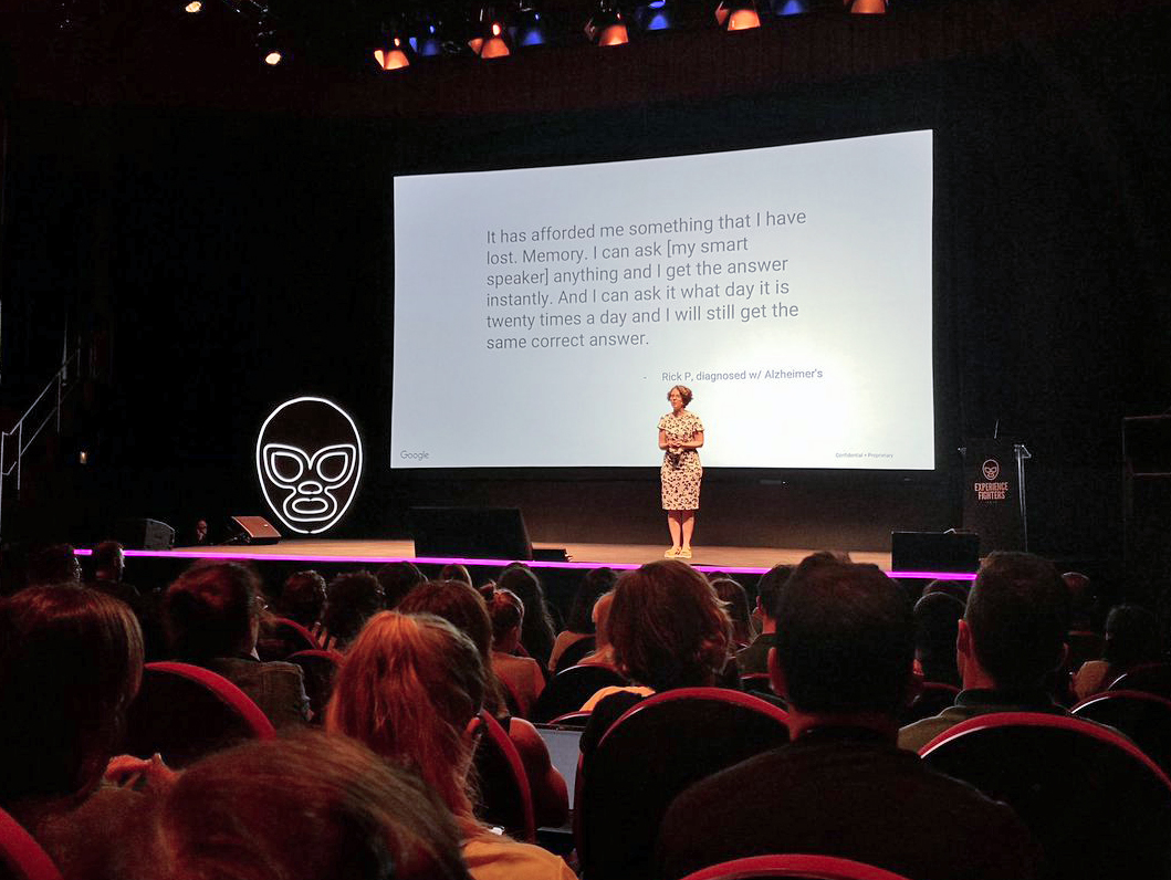 Cathy Pearl presenting on a sleek stage with a large slide screen behind her.