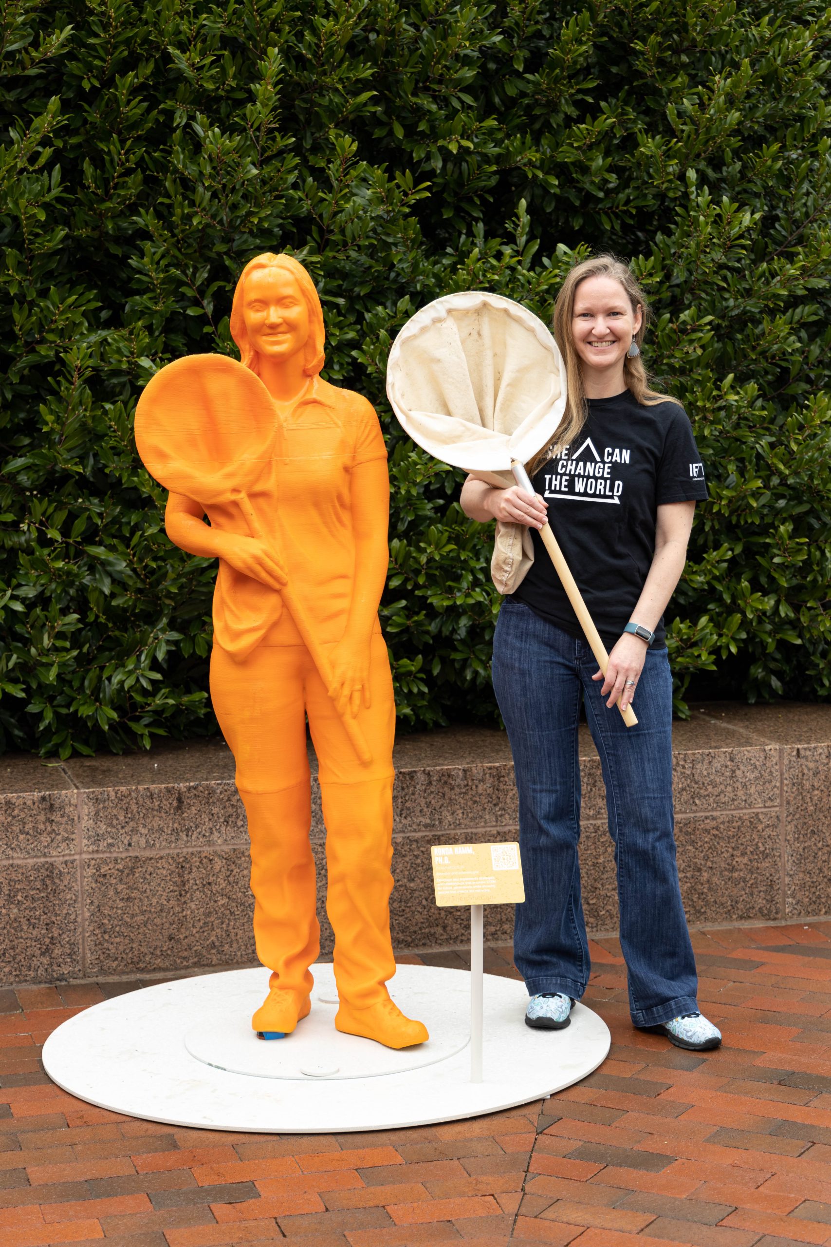 Dr. Ronda Hamm stands next to her statue, smiling and holding an insect net just like her statue.