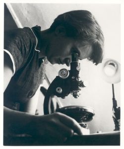 Black and white photo of Rosalind Franklin looking into a microscope