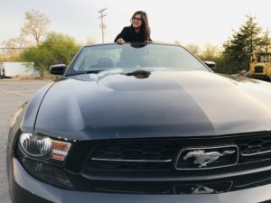 Shrishti is standing in a Mustang convertible, posing with her crossed arms resting on the top of the windshield.