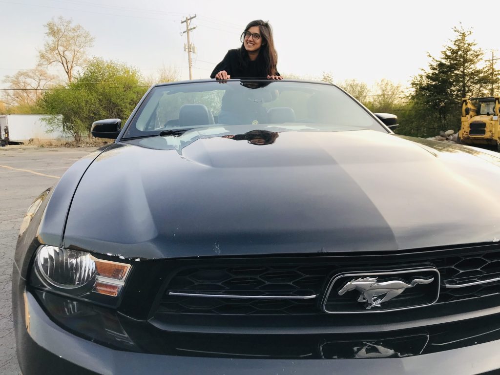 Shrishti is standing in a Mustang convertible, posing with her crossed arms resting on the top of the windshield.