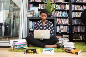 A Black woman sits on the floor with her laptop surrounded by colorful books on coding and a green plant behind her.