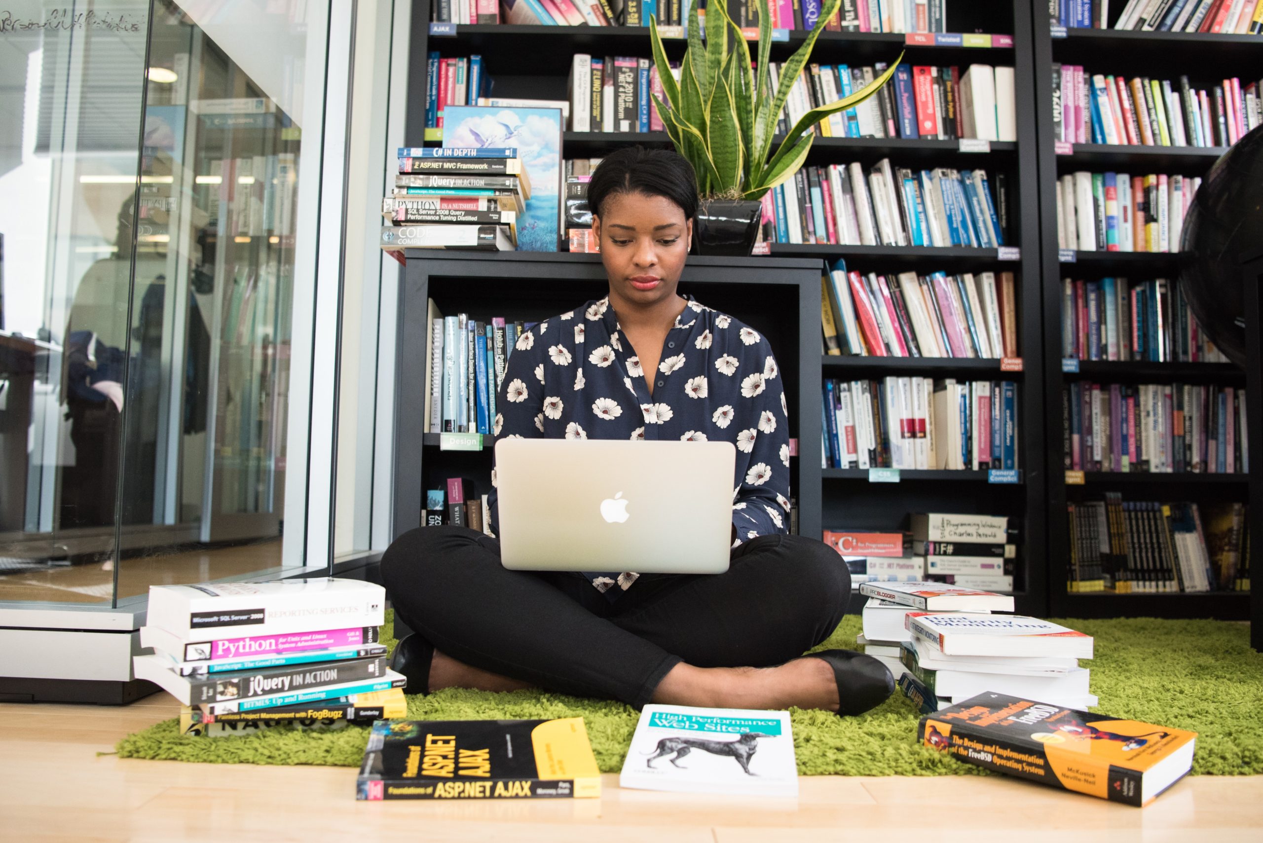 A Black woman sits on the floor with her laptop surrounded by colorful books on coding and a green plant behind her.